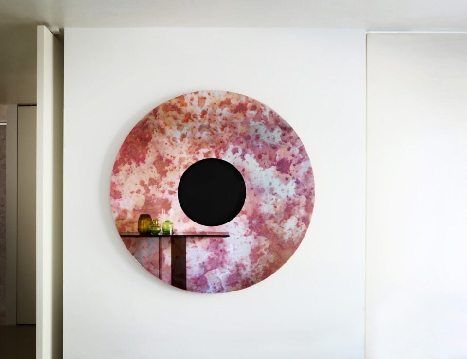 Round glass mirror with pink and red speckled finish mounted on wall.