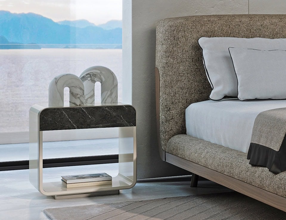 Lacquered wood bedside table with open shelf beside upholstered bed.